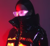 Person wearing reflective futuristic glasses and a shiny black puffer jacket under red and purple lighting.